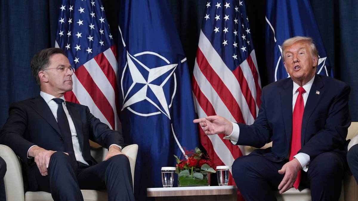 Dwóch mężczyzn siedzi przed flagami USA i NATO, jeden z nich gestykuluje.