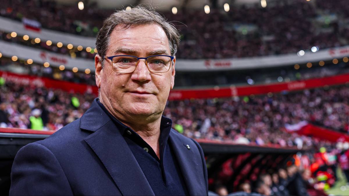 Trener piłkarski Jan Urban w okularach, ubrany w ciemny garnitur, stoi na tle trybun stadionu wypełnionych kibicami.