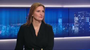 Kobieta w czarnej marynarce siedzi w studiu telewizyjnym, z poważnym wyrazem twarzy, na tle ekran&oacute;w wyświetlających nocny widok miasta.