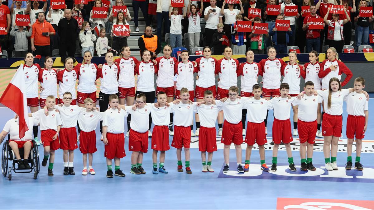 Drużyna piłkarki ręcznej w czerwonych strojach stoi na boisku, trzymając flagę Polski, z publicznością w tle.