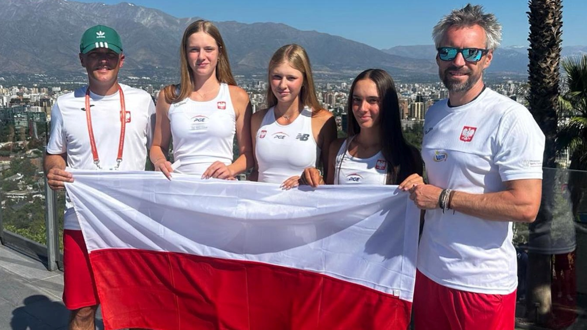 Pięć osób, w tym cztery młode tenisistki i jeden mężczyzna, pozuje z biało-czerwoną flagą Polski na tle miasta.