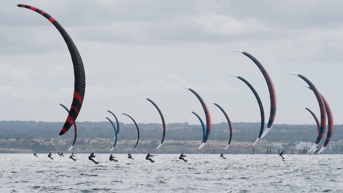Grupa kitesurfer&oacute;w na wodzie, każdy z kolorowym żaglem, na tle chmur i linii brzegowej.