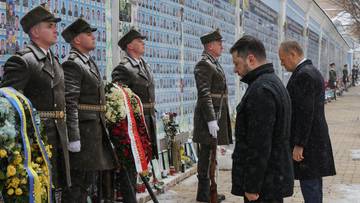 Przedstawiciele władz i żołnierze stojący przed ścianą z portretami upamiętniającymi ofiary konfliktu.