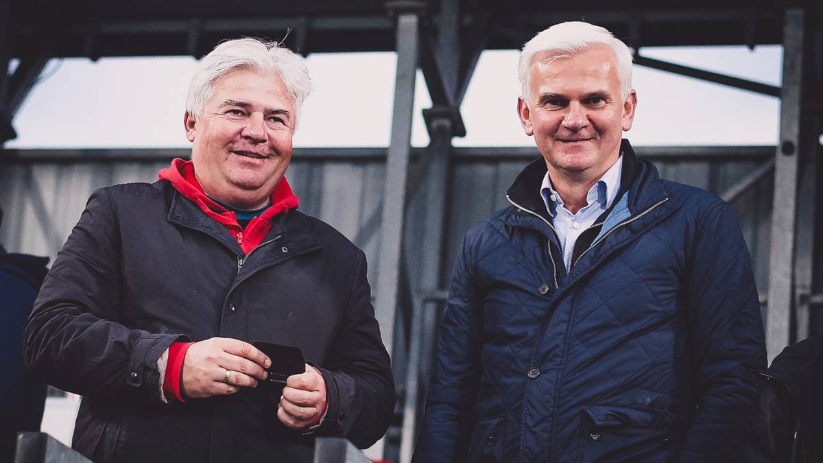 Dwóch mężczyzn w średnim wieku, obaj z siwymi włosami, stoją obok siebie na zewnątrz, patrząc w obiektyw z delikatnym uśmiechem. Obaj są ubrani w kurtki.