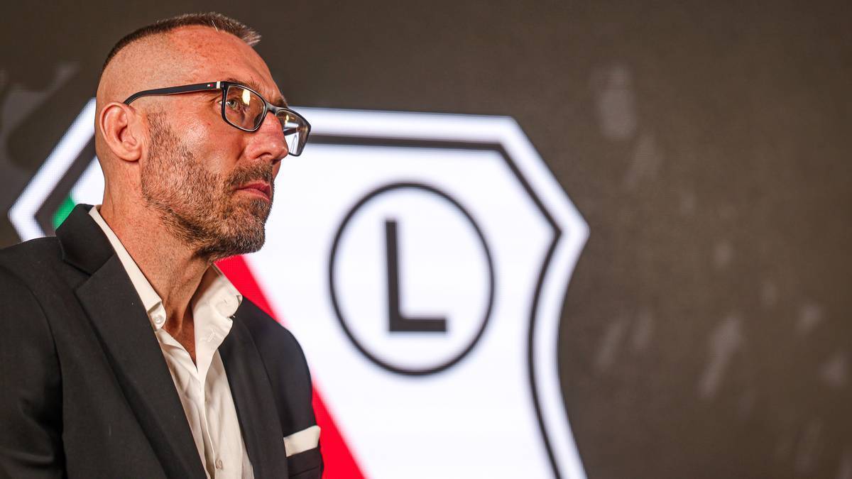 Mężczyzna w okularach i marynarce z widocznym zarysowanym logo "L" w tle.