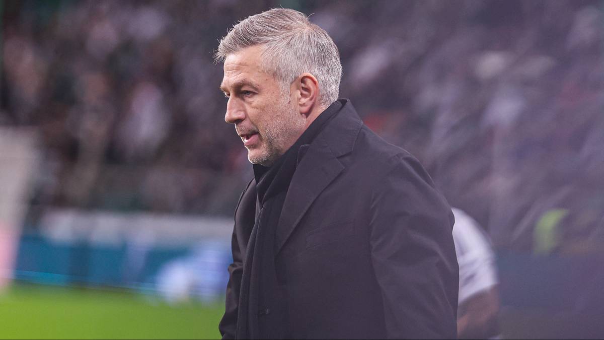 Mężczyzna w średnim wieku z siwymi włosami i ciemnym płaszczem, patrzący w lewo, na tle trybun stadionu.