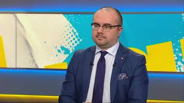 Mężczyzna w okularach i granatowym garniturze, z białym kołnierzykiem i fioletowym krawatem, siedzi przed ekranem z abstrakcyjnym wzorem w tle.