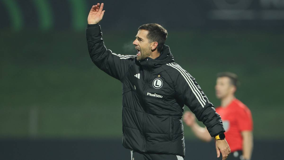 Trener Legii Warszawa w kurtce z logo klubu podnosi rękę, wydając polecenia.