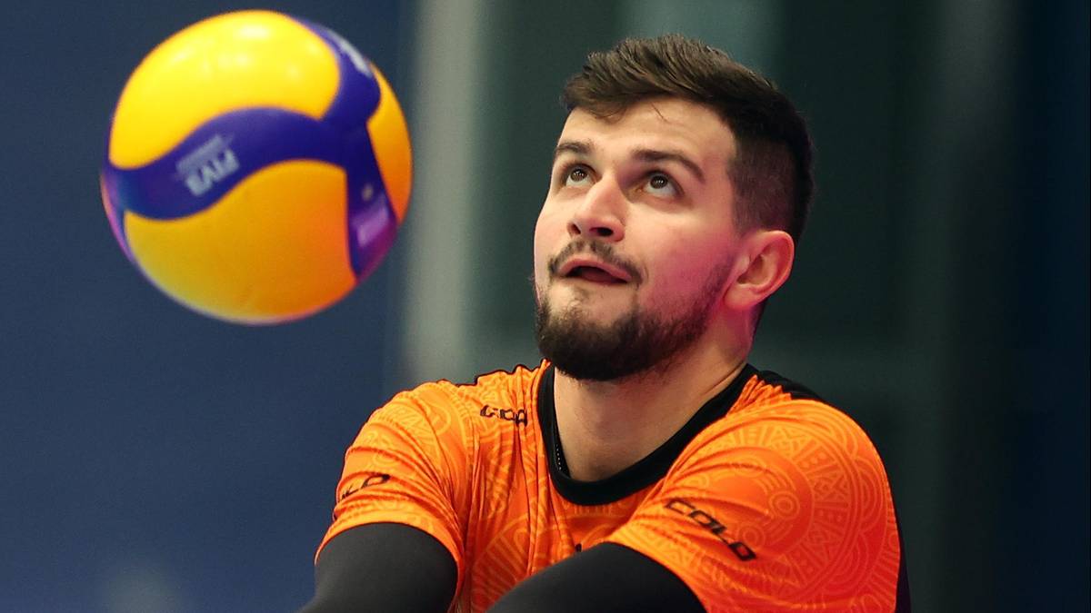 Mężczyzna w pomarańczowej koszulce patrzy w górę na lecącą piłkę do siatkówki.