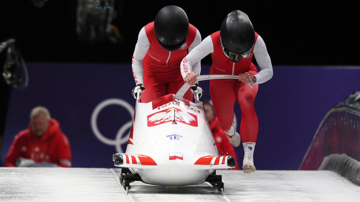 Dwie zawodniczki w czerwonych strojach z białymi elementami i kaskami wciskają bobslej na torze lodowym.