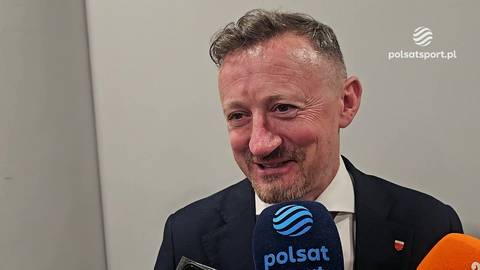 Adam Małysz uśmiecha się, trzymając mikrofon Polsat Sport przed twarzą.