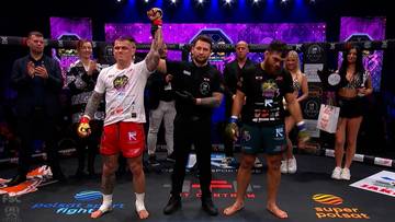 Dwóch mężczyzn w strojach sportowych unosi ręce w geście zwycięstwa na tle sceny z zawodnikami i osobami z obsługi gali.