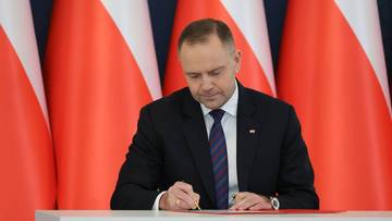 Prezydent podpisuje dokumenty na tle flag Polski.