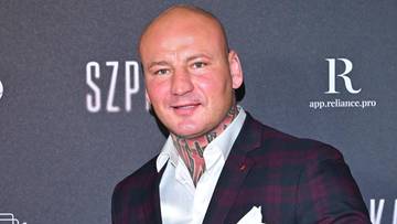 Łysy mężczyzna w garniturze w kratę i białej koszuli uśmiecha się do aparatu, zaciskając prawą pięść.