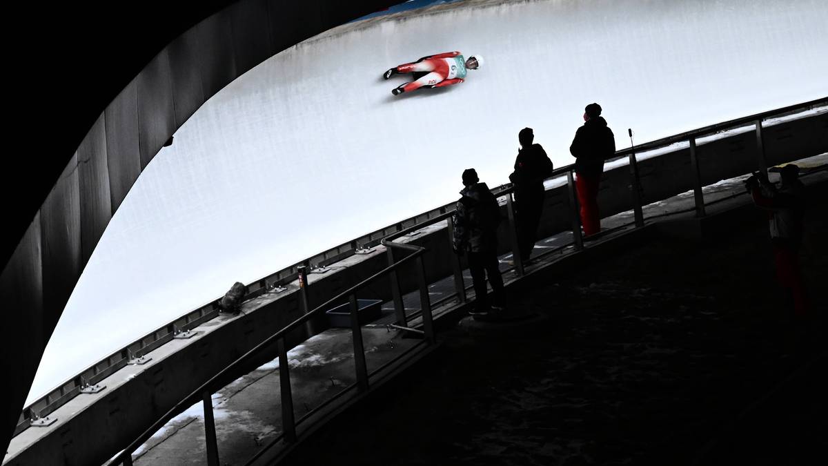 Zawodnik w stroju narciarskim leżący na sankach, zjeżdża po śliskim torze lodowym. Obok toru stoją obserwatorzy.