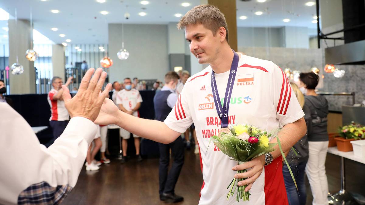 Siatkarz w koszulce z flagą Polski i napisem "BRA WO MIAŁ" odbiera bukiet kwiatów od innej osoby.