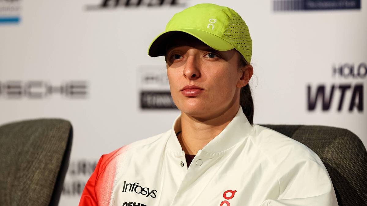 Iga Świątek siedzi na krześle, ubrana w białą bluzę z czerwonymi i białymi elementami. Na głowie ma limonkową czapkę z daszkiem. Jej wyraz twarzy jest poważny i skupiony.