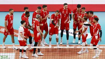 Grupa męskich siatkarzy w czerwonych strojach sportowych na boisku, przygotowujących się do meczu. Widoczni są zawodnicy z numerami na koszulkach.