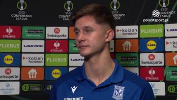 Młody mężczyzna w niebieskiej koszulce sportowej stoi przed ścianą z logo sponsor&oacute;w.