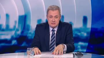 Mężczyzna w garniturze i krawacie siedzi przy stole w studio telewizyjnym, przed nim szklanka z wodą i dokumenty.