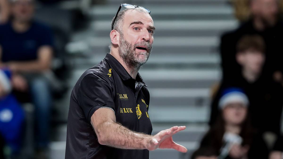 Trener siatkówki w czarnej koszulce z żółtym logo, gestykulujący podczas meczu lub treningu.