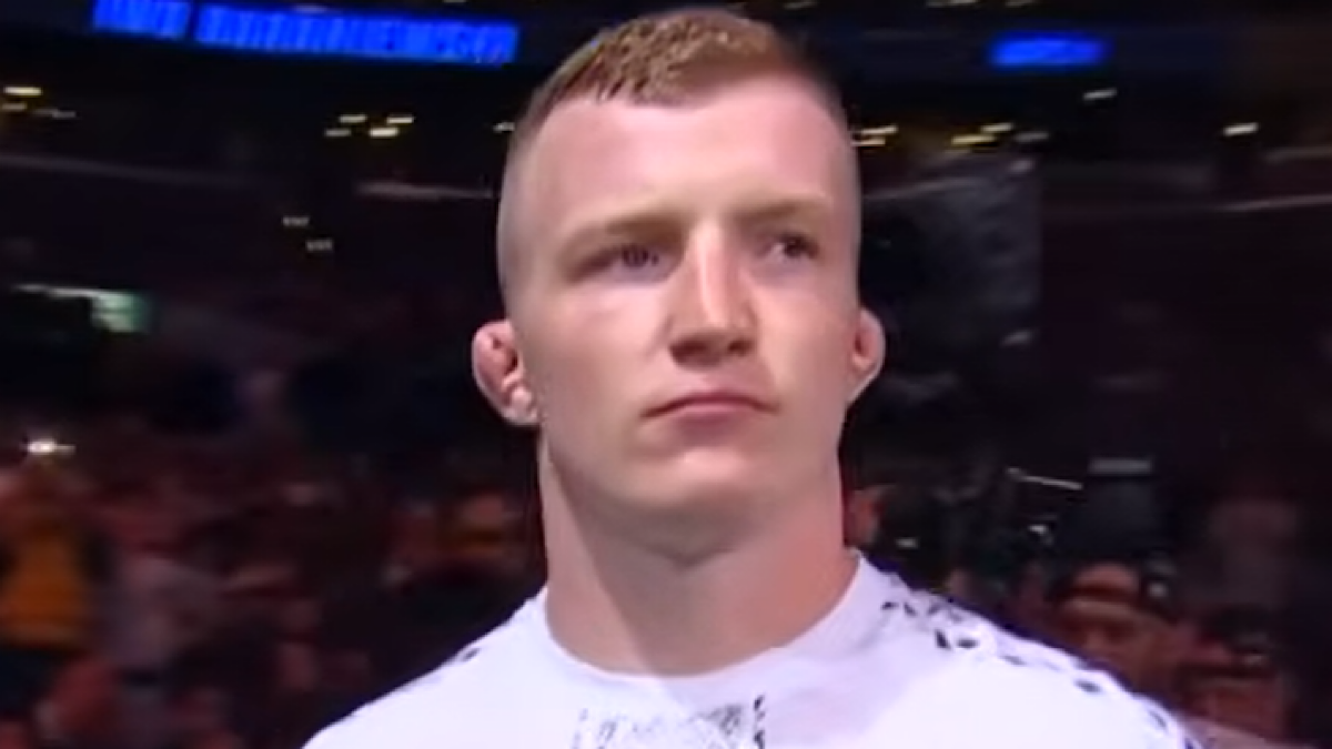 Młody biały zawodnik sport&oacute;w walki w białej koszulce z kr&oacute;tkimi blond włosami i poważnym wyrazem twarzy, na tle rozmazanej publiczności.