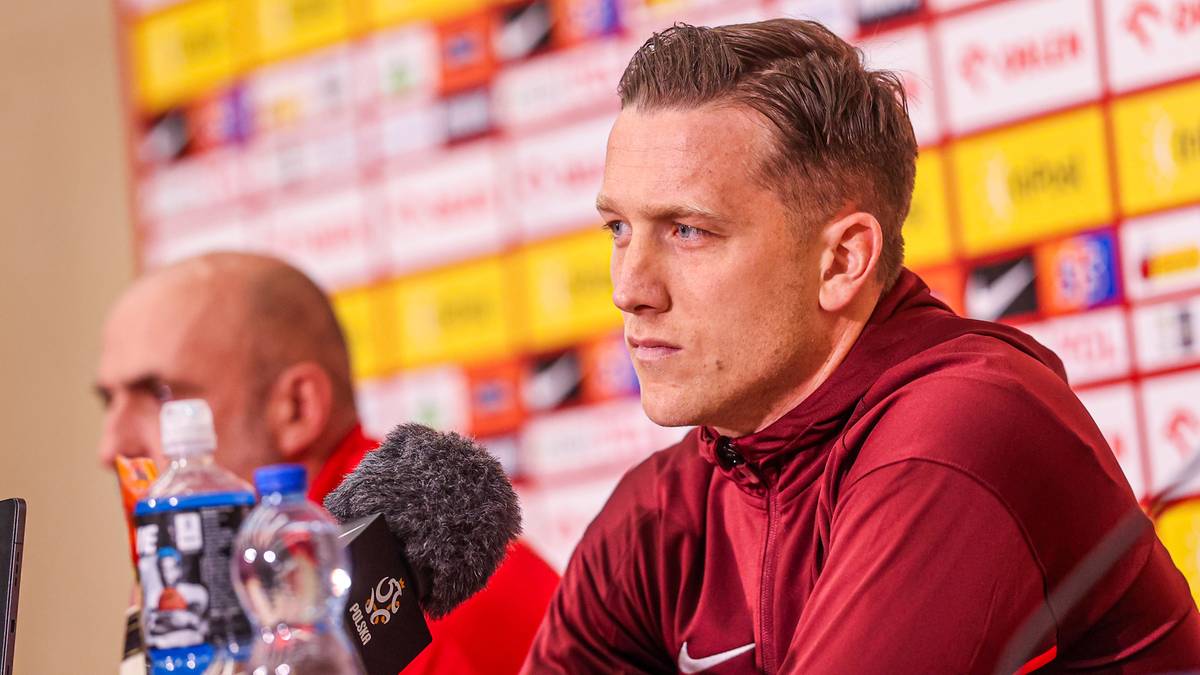 Mężczyzna w czerwonej bluzie z kapturem patrzy w bok z poważną miną. Za nim siedzi inny mężczyzna, a przed nimi stoją butelki z wodą i mikrofon.