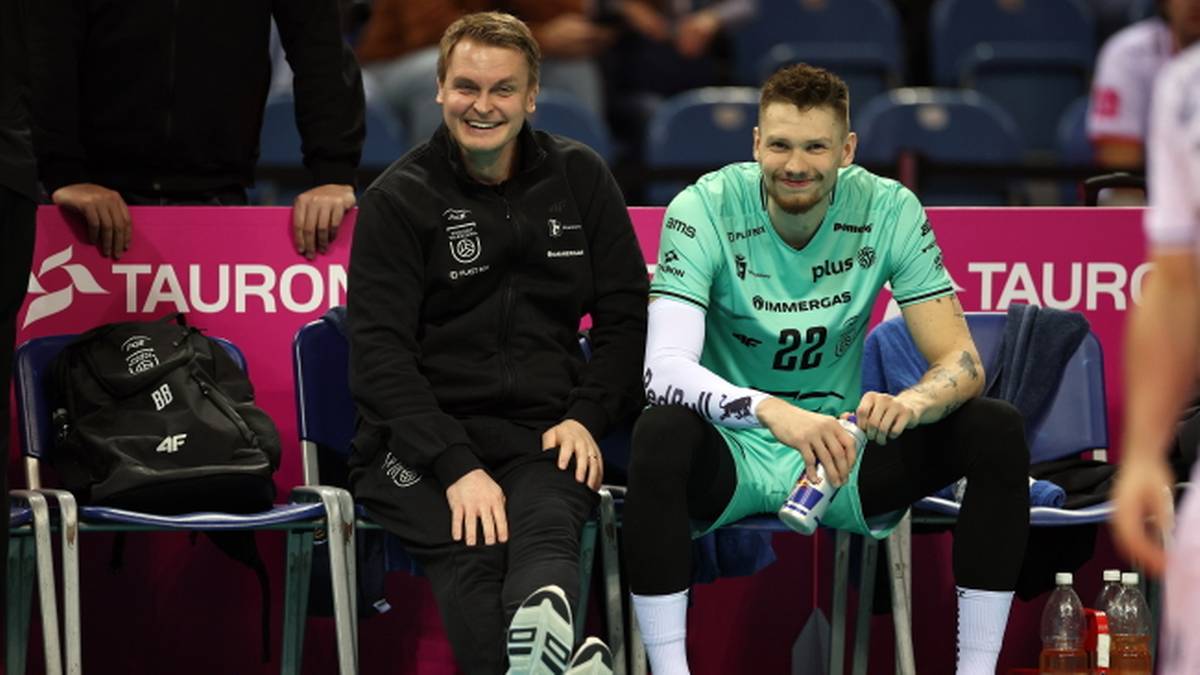 Dwóch uśmiechniętych siatkarzy siedzi obok siebie na ławce rezerwowych.