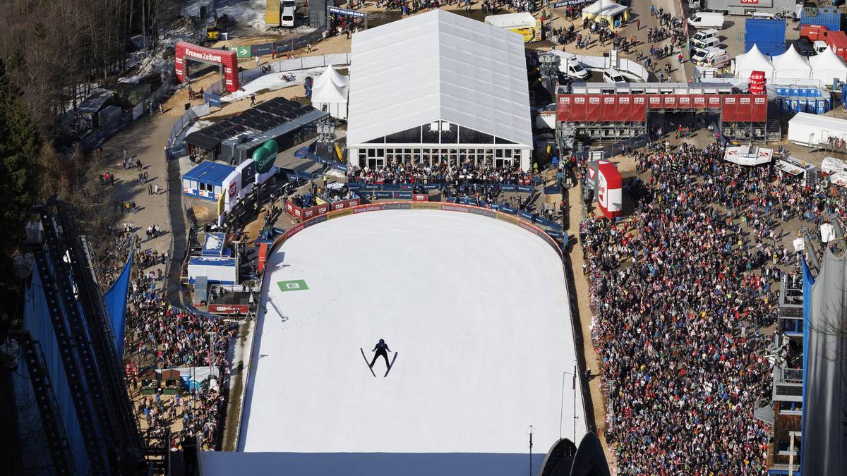 Widok z g&oacute;ry na skocznię narciarską z zawodnikiem na zeskoczni i pełnymi trybunami.