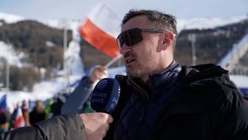 Adam Małysz w okularach przeciwsłonecznych i czapce, trzymający mikrofon, z polską flagą w tle.