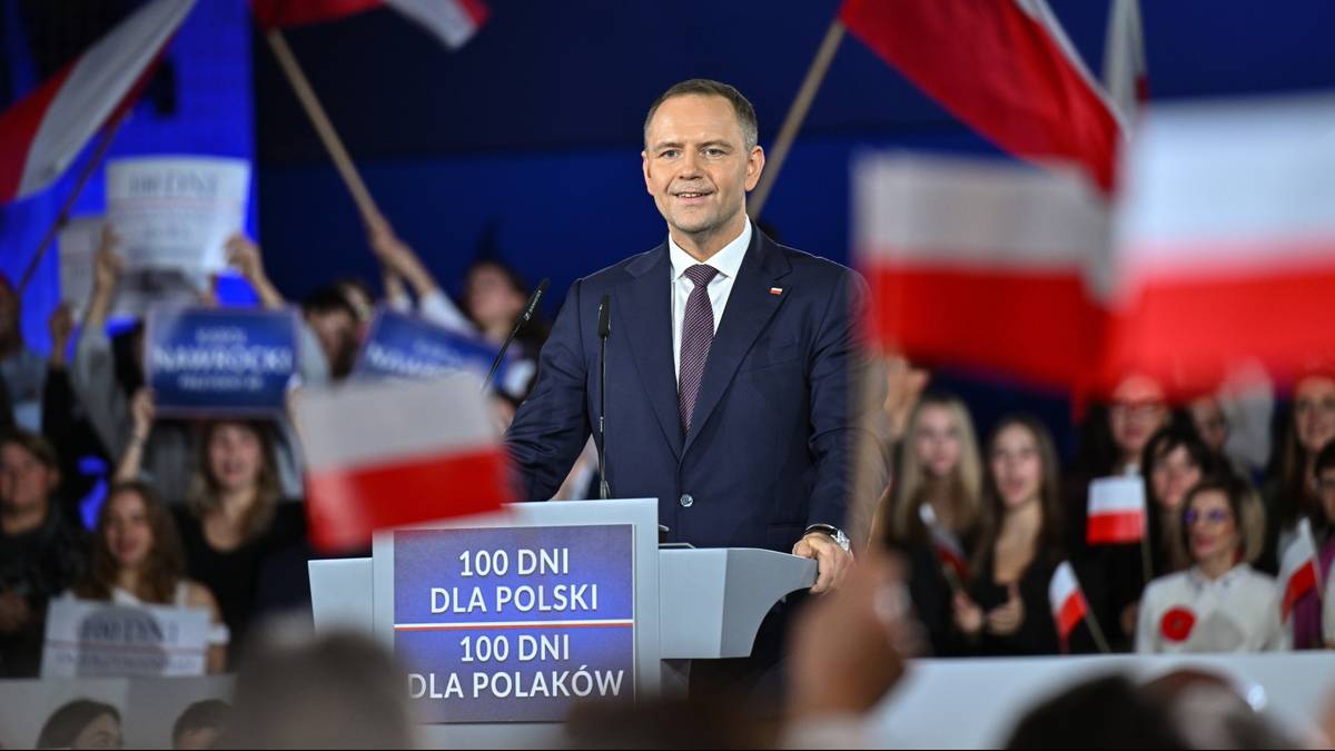 Mężczyzna w garniturze za mównicą z tablicą "100 DNI DLA POLSKI" na tle flag Polski i tłumu widzów.