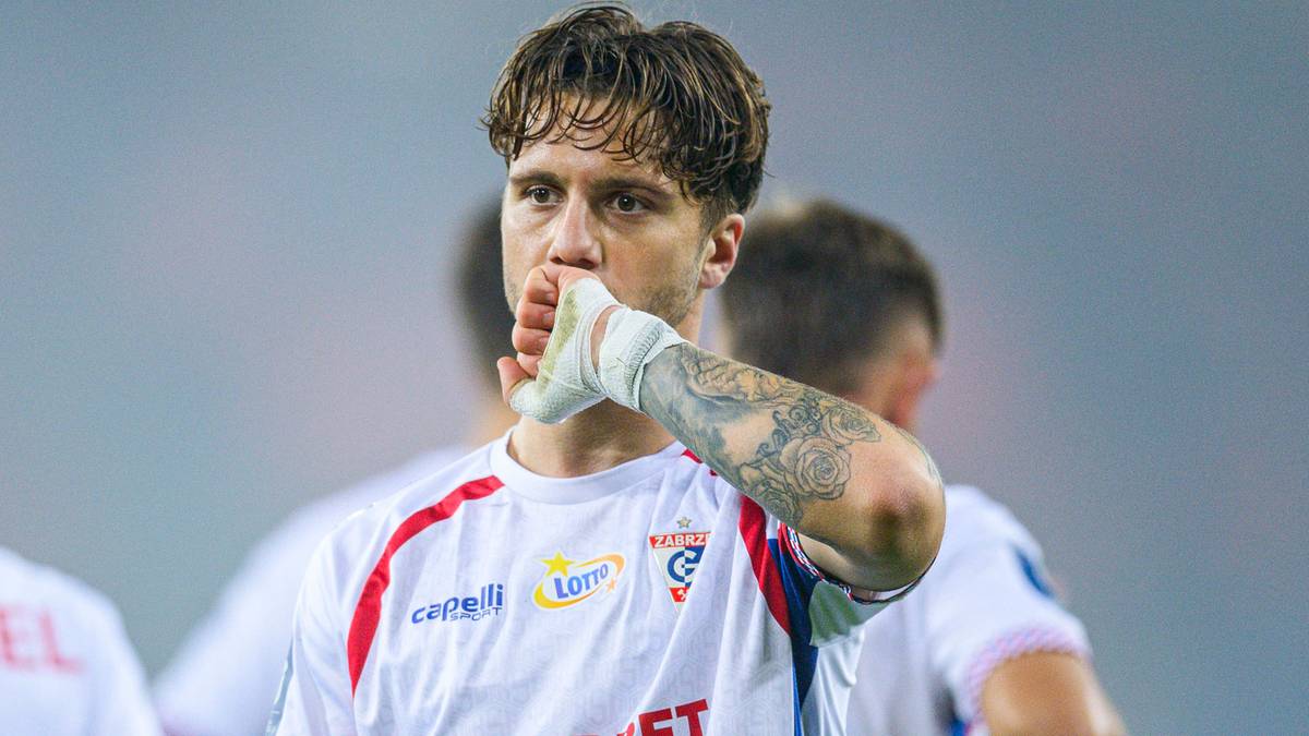 Piłkarz Górnika Zabrze w białej koszulce, z bandażem na dłoni zakrywającej usta, gestykuluje.