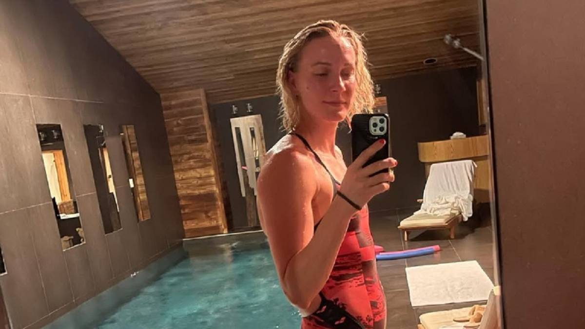 Kobieta w czerwonym stroju kąpielowym robi sobie zdjęcie z telefonem w ręku, stojąc obok basenu. Jest w ciąży.