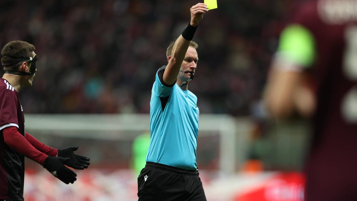 Sędzia trzymający żółtą kartkę w górze na boisku piłkarskim. Po lewej stronie obrazu stoi piłkarz w burgundowej koszulce.