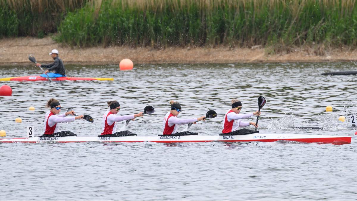 Cztery zawodniczki w kajaku siedzą obok siebie, wiosłując jednocześnie. Wszystkie mają na sobie białe koszulki z czarnymi rękawami i czerwonymi pasami, a także czarne spodenki. Na głowach mają czarne czepki.
