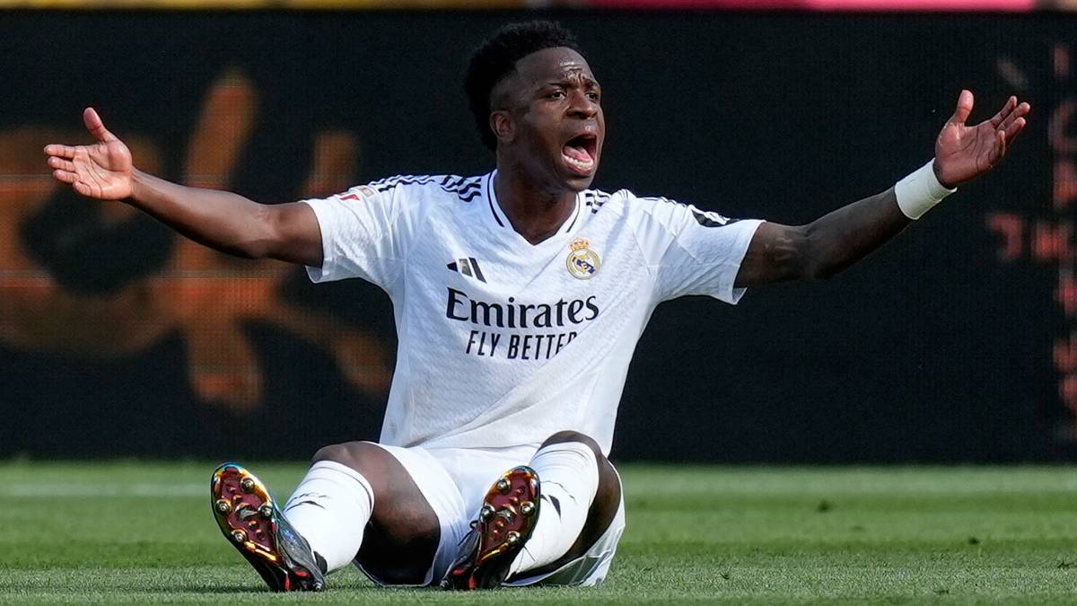 Piłkarz Realu Madryt, Vinicius Junior, siedzi na trawie z otwartymi rękami i wyrazem rozpaczy na twarzy, ubrany w białą koszulkę.