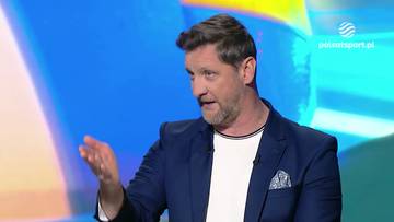 Piotr Gruszka, mężczyzna w średnim wieku, w granatowej marynarce i białej koszulce, gestykuluje prawą ręką podczas wypowiedzi na tle kolorowego tła.