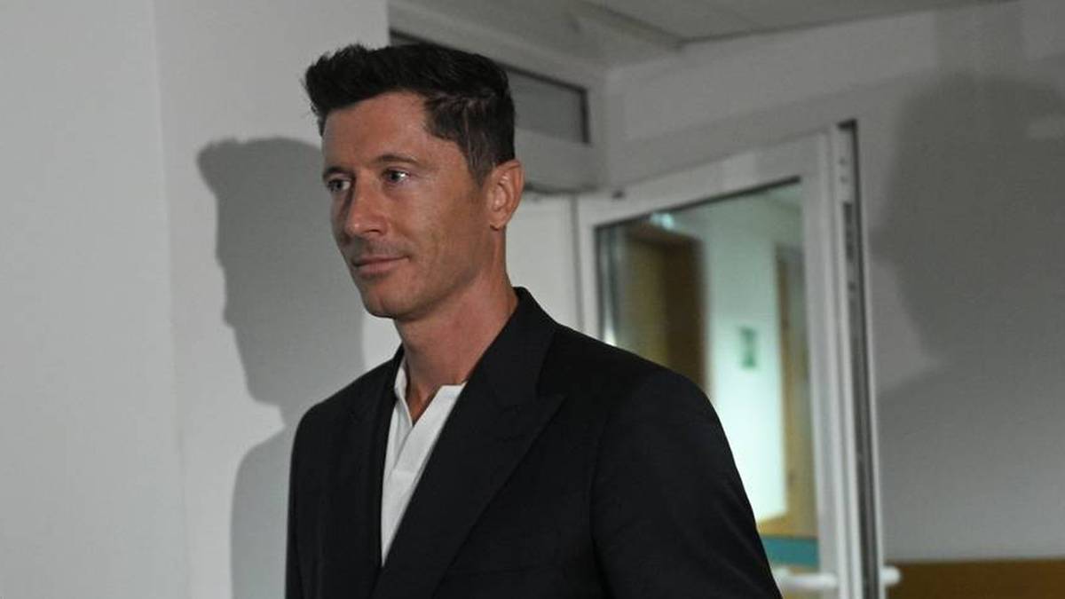 Portret Roberta Lewandowskiego, ubranego w ciemną marynarkę i białą koszulę, stojącego wewnątrz pomieszczenia z drzwiami w tle.