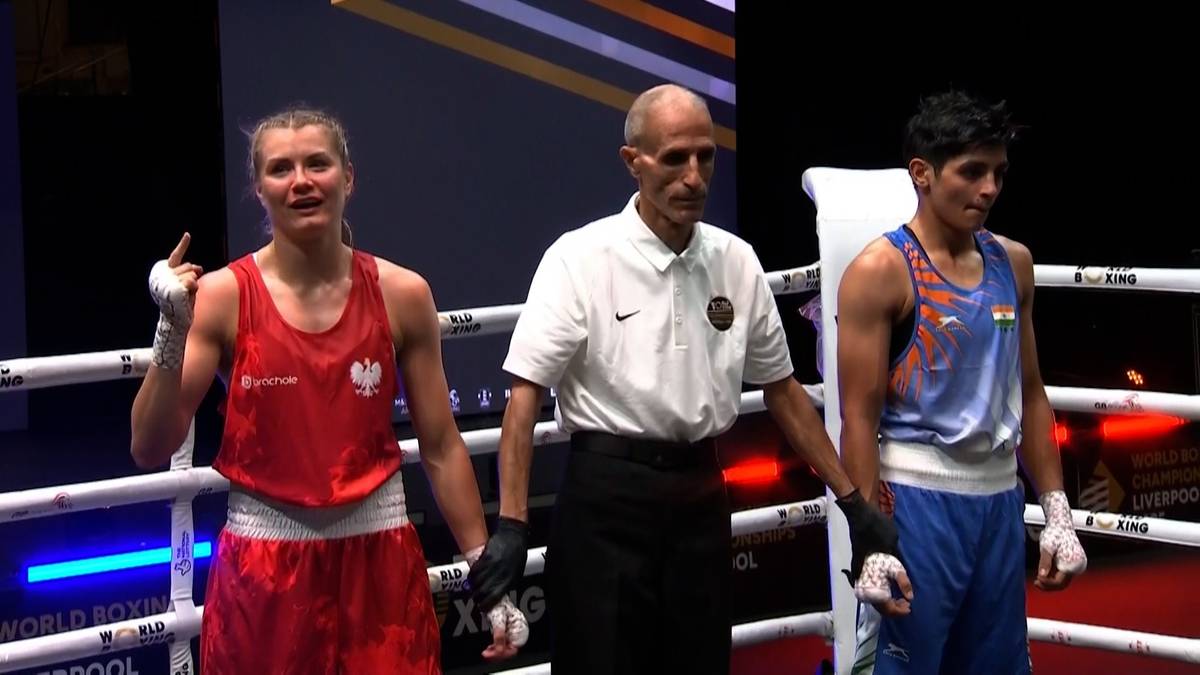 Młoda kobieta w czerwonym stroju bokserskim, z biało-czerwoną flagą na piersi, stoi na ringu bokserskim z podniesioną ręką w geście zwycięstwa. Obok niej stoi sędzia w białej koszuli, a po drugiej stronie młody mężczyzna w niebieskim stroju bokserskim.