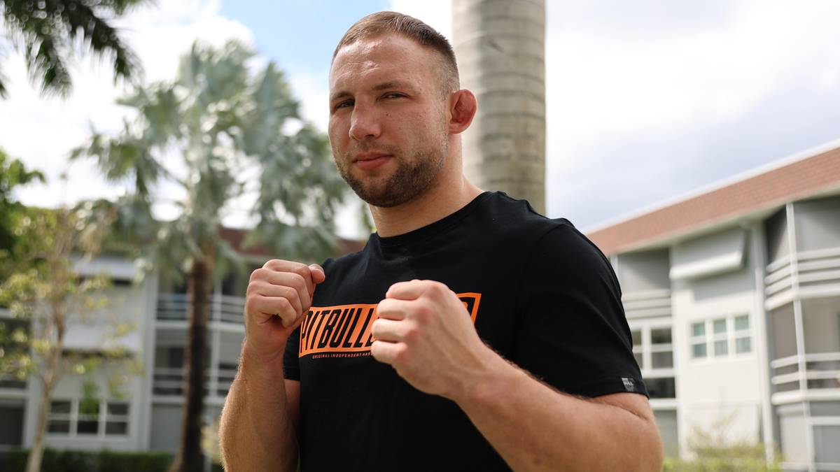 Mężczyzna w czarnej koszulce z pomarańczowym napisem, stoi na zewnątrz z zaciśniętymi pięściami.