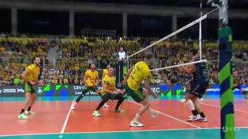 Gracz w ż&oacute;łtej koszulce wykonuje atak w meczu siatk&oacute;wki, podczas gdy przeciwnicy w ciemnych strojach pr&oacute;bują go zablokować.