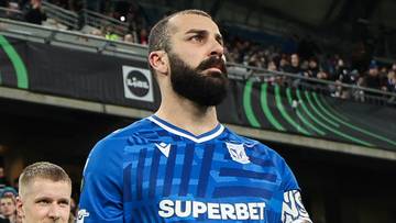 Piłkarz w niebieskiej koszulce prowadzi grupę chłopców w żółto-niebieskich strojach na boisko, w tle widać trybuny wypełnione kibicami.