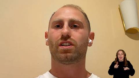 Mężczyzna z brodą i słuchawkami bezprzewodowymi na uszach m&oacute;wi, w tle widoczna osoba migająca.