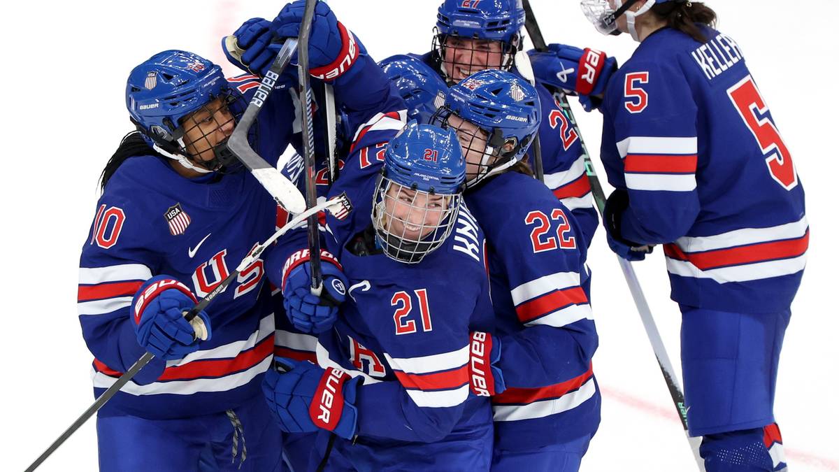 Grupa hokeistek USA w niebieskich strojach świętuje zwycięstwo, niektóre trzymają kije hokejowe.