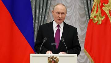 Władimir Putin stoi za mównicą, przemawiając w trakcie uroczystości. Za nim widoczne są flagi Rosji.