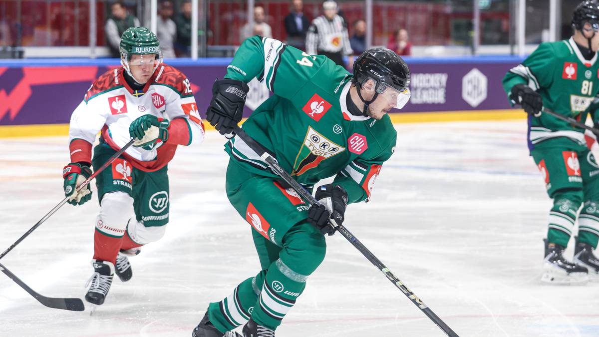 Hokeista GKS Tychy w zielonym stroju prowadzi krążek na lodowisku, ścigany przez przeciwnika.