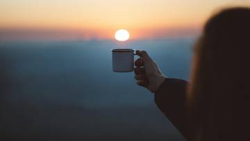 Dłoń trzymająca kubek na tle zachodzącego słońca.