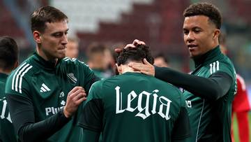 Piłkarze Legii Warszawa w zielonych strojach treningowych na boisku piłkarskim, jeden zawodnik poprawia włosy drugiemu.