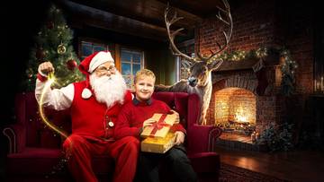 Święty Mikołaj w czerwonym stroju siedzi na czerwonej kanapie obok chłopca w czerwonym swetrze. Chłopiec trzyma prezent. Za nimi znajduje się kominek z ogniem i choinka.