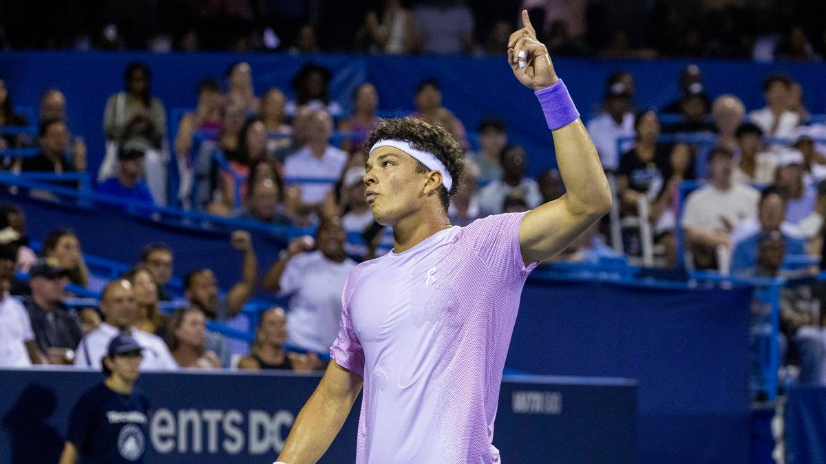 Tenisista w koszulce z krótkim rękawem i opaską na czole, podnosi prawą rękę do góry.